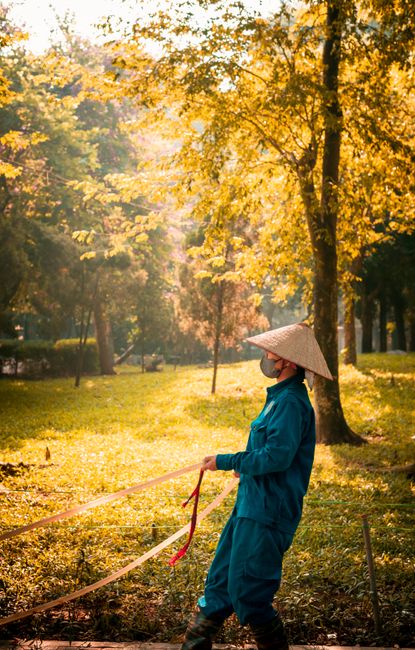 A gardener wearing a traditional conical straw hat, blue jacket, and black boots is working in a park during autumn, holding a gardening tool with a red strap. The scene features a lush green lawn bor