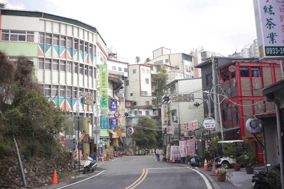 A street scene in an urban area with a gentle curve in the road, flanked by multi-story buildings of various architectural styles and colors. On the left, a modern round building with a green and oran