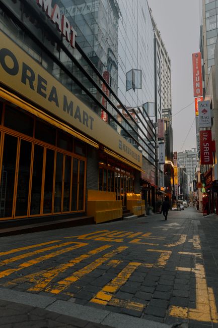 A street scene in an urban area with a gentle curve in the road, flanked by multi-story buildings of various architectural styles and colors. On the left, a modern round building with a green and oran